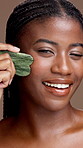 Face, gua sha and massage with black woman in studio for natural skin treatment or wellness. Facial skincare, jade stone and smile of model person on brown background for cosmetic dermatology routine