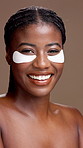 Face, skincare and black woman with eye mask for beauty, glow or health. Portrait, cosmetics and smile with collagen patches for dermatology, shine and reduce dark circles on brown studio background