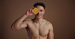Face, man and orange for skincare in studio for vitamin c, brighten skin and natural detox. Portrait, male person and fruit with beauty, organic benefits and dermatology exfoliate on brown background