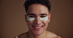 Face, eye patches and man with dermatology, smile and skincare on brown studio background. Portrait, space and happy person with cosmetics, spa wellness and grooming with healthy skin and laughing