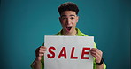 Shocked, sale and face of man in studio with poster for discount, bargain and deal news on blue background. Happy, space and portrait of person with banner for announcement, promotion and information