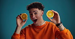 Face, surprise or man in studio with orange, smile or citrus isolated on blue background. Play, happy or shocked model with fruit for healthy diet, nutrition or wellness with vitamin c or benefits