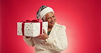 Christmas, gift and shake with face of old woman in studio for announcement, holiday season and laugh. Festive celebration, Xmas present and December with elderly person on red background space