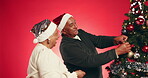 Old couple, Christmas and decorating tree in studio, festive love with bauble or ornament for holiday decor. Help, celebration and senior people with glitter ball for December break on red background