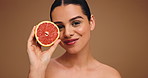 Face, woman and grapefruit for skincare in studio for vitamin c, anti aging benefits and hydration. Smile, person and fruit for beauty, organic ingredients and natural facial glow on brown background