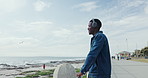 Happy, thinking and man with headphones by ocean for listening to audio, podcast and music. Relax, beach and person on promenade for streaming playlist, radio and song on holiday, weekend or travel