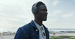 Happy, music and black man with headphones by ocean for listening to audio, podcast and playlist. Relax, beach and person on promenade for streaming, radio and song on holiday, weekend and vacation