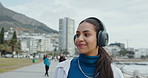 Happy, walking and woman with headphones by ocean for listening to audio, podcast and music. Relax, city and person on promenade for streaming playlist, radio and song on holiday, weekend or travel