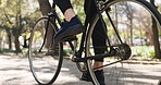 Person, legs and cycling with bicycle in park for travel, commute or eco friendly transportation. Shoes, cyclist and ride with bike pedals on road for carbon capture, fitness or exercise in nature