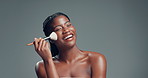 Funny, brush and black woman in studio, makeup and foundation on gray background. Space, cosmetic tool and African person with shine, laughing and dermatology with glow, beauty and aesthetic