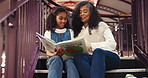 Book, library and grandmother reading with child for bonding, connection or development. Happy, education and senior woman with girl kid for story together on stairs for knowledge and learning.