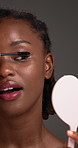 Black woman, eye lashes and mirror with mascara in studio for beauty makeup on a gray background. Female person, model or cosmetics with curls, wand or stick for thickness, darker volume or texture