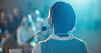 Woman, back and podium with crowd in auditorium for presentation or speech in business seminar. Female person, employee or speaker with microphone or audience for workshop or corporate trade show