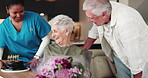 Senior woman, birthday and cake with flowers at house for party, celebration and surprise. Elderly couple, bouquet and gift with nurse for special occasion, sweet treat and romantic kiss of milestone