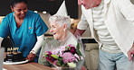 Senior woman, birthday and cake with flowers in home for party, celebration and surprise. Elderly couple, bouquet and gift with nurse for special occasion, sweet treat and romantic kiss of milestone