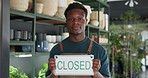 Happy, closed sign and face of black man in store for sustainability, plants or greenery at small business. Board, smile and portrait of African botanist with announcement at at eco friendly nursery.