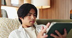 House, tablet and man on couch, typing and relax with digital app for online subscription. Apartment, tech and Asian person on sofa, internet and reading for news report, connection and daily updates
