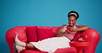 Phone, relax and black woman on sofa in studio with networking, social media or communication on mobile app. Happy, contact and African female person with cellphone for connection by blue background.