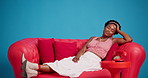 Tired, stretching and black woman on couch in studio, chilling and comfortable for weekend peace. Relaxation, fatigue and person resting on sofa with smile for day off, wellness and blue background