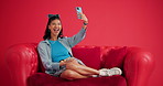 Selfie, cellphone and happy woman on sofa in studio, relax and social media to capture moment. Mobile user, smile and person on couch, profile picture and app for photography on red background