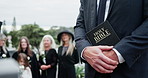 Hands, bible and priest at cemetery for funeral, service and crowd with Christianity for mourning. People, pastor or reverend with family, holy scripture and sermon for burial ceremony at graveyard