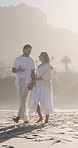 Couple, walking and holding hands on beach with holiday, conversation and laugh for bonding together. Happy, married people and outdoor by ocean with love, summer vacation and discussion for travel.
