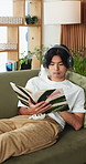 Headphones, reading and man with novel on sofa listening to music, playlist or album in living room. Audio tech, relax and Asian person with story, book or literature with streaming podcast in home.
