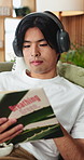 Headphones, reading and man with book on sofa listening to music, playlist or album in living room. Audio tech, relax and Asian person with story, novel or literature with streaming podcast in home.