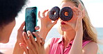 Outdoor, friends and donut eyes for photography, playful social media post and funny holiday memory. Vacation, phone or happy women with dessert for silly picture, tongue out or laugh for beach trip
