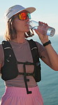Sea, woman and water bottle on hiking break to relax, rest and ocean breeze with thirst. Female person, fitness and trekking outdoor with fresh drink, sunglasses and smile for exercise and health