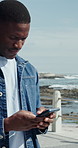 Ocean, walking and black man with phone, scroll for online chatting and app for tourism. Beach, connection and person with tech for social media, network and vacation with communication in Miami