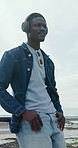 Headphones, outdoor and black man by beach for holiday with listening to music, playlist or radio. Relax, audio tech and person on promenade by ocean for streaming album on vacation or weekend trip.