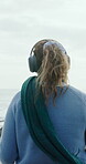 Beach, woman and relax with headphones, outdoor and listening to music on winter holiday and online. Person, back and streaming audio with tech on weekend adventure, ocean and sound on vacation