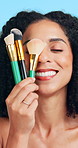 Happy, brushes and woman with makeup for shine, cosmetics and skincare on blue studio background. Tools, facial foundation and person with aesthetic, glowing and product for beauty and dermatology