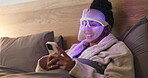 Blue light, mask and woman with phone for skincare, relax or wellness with antibacterial facial. Health, beauty tech and female person on bed with LED phototherapy for dermatology treatment in home