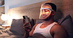 Red light, mask and man with phone for skincare, relax or wellness with collagen production. Health, beauty tech and male person on bed with LED phototherapy for dermatology treatment in home