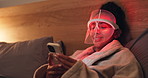 Red light, mask and woman with phone for skincare, relax or wellness with collagen production. Health, beauty tech and female person on bed with LED phototherapy for dermatology treatment in home