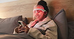 Red light, mask and woman with phone for dermatology, relax or wellness with collagen production. Health, beauty tech and female person on bed with LED phototherapy for skincare treatment in home