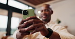Hands, black man and relax in home with phone, social media and browsing website in living room. Smile, male person or reading at house with mobile, communication and download ebook for weekend break