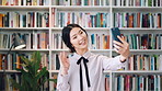 Phone, video call and Asian woman in library for education, course update and wave in university. Mobile, elearning and student on virtual chat for curriculum info, greeting and smile by bookshelf