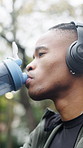 Training, break and black man drinking water in park with headphones, workout and fitness hydration. Runner, tired and athlete with bottle of h2o in nature for sports recovery, exercise and nutrition