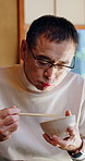 Asian man, eating and food at house with chopsticks, healthy rice and traditional meal for lunch. Mature person, bowl and utensils in lounge for brunch, nutrition snack and authentic Chinese cuisine 