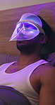 Mask, skincare and man with red light therapy on bed for relax, beauty or facial wellness. Health, cosmetic and person with face LED treatment for collagen production in bedroom at apartment.