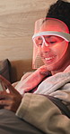 Mask, cellphone and woman with red light therapy on bed for skincare, beauty or facial wellness. Happy, phone and person with face LED treatment for collagen production in bedroom at apartment.