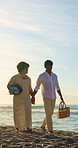 Couple, holding hands and chat with picnic basket at beach, bonding and summer holiday with partner. Outdoor, happy people and walk with spouse on coastal vacation, space and commitment in nature