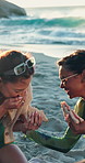 Friends, laugh and sandwich by ocean with summer holiday, funny story or bonding together on weekend. Happy, people and eating food outdoor at beach with hangout, gossip or humor on tropical vacation