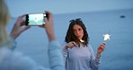 Sparklers, beach and friends with phone taking pictures for happy memory. Fireworks, sunset and girls or women at seashore taking a photo on 5g mobile smartphone for profile picture on social media.