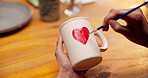 Pottery, painting and hands with cup in&nbsp;workshop, creative and drawing heart shape with paintbrush. Artisan, craftsmanship and person with handmade mug, above and art process with equipment or color